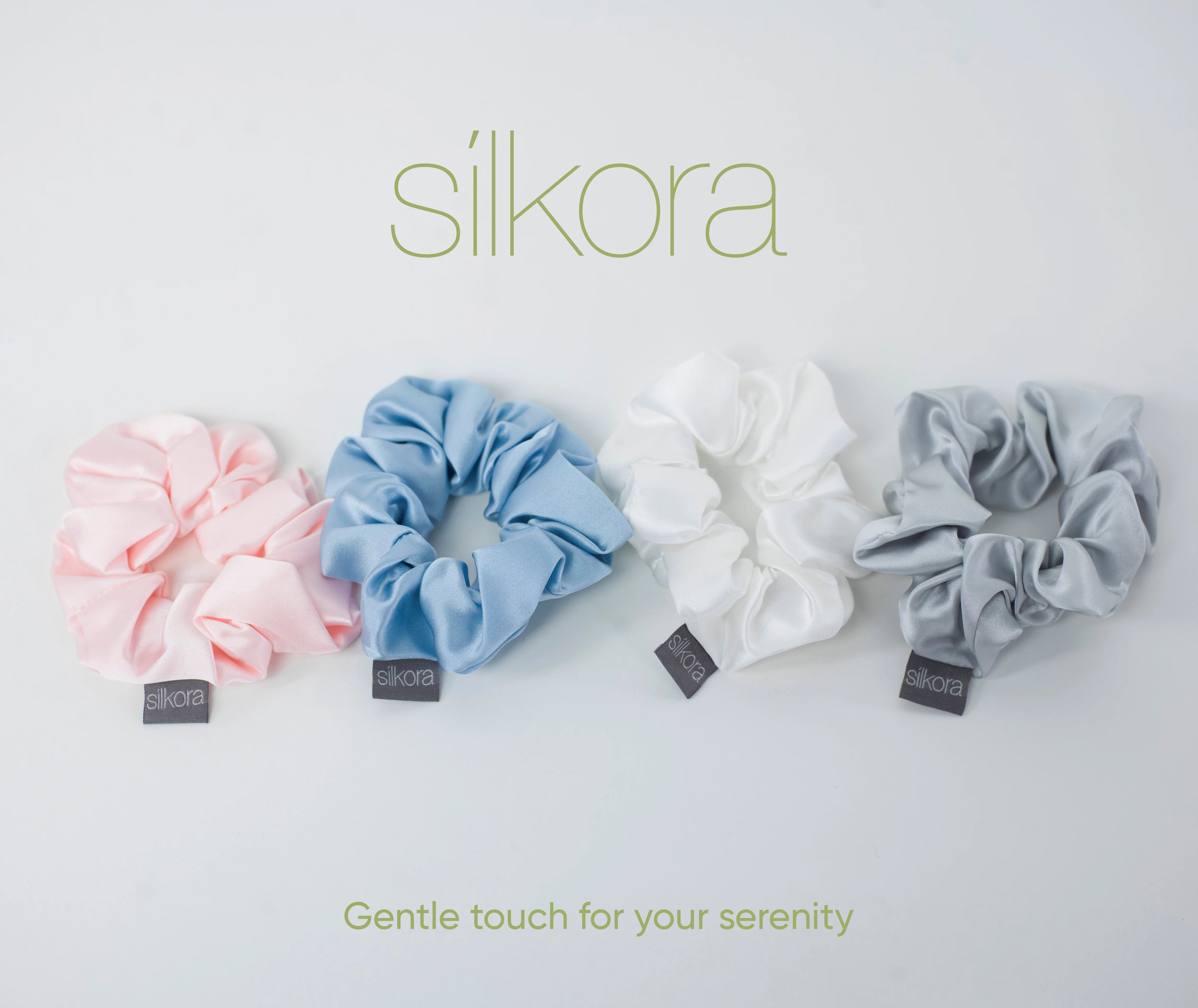 Vier grote Silkora scrunchies in lotus roze, icy blauw, wit en zilvergrijs op een lichte achtergrond met het Silkora-logo en de tekst "Gentle touch for your serenity".